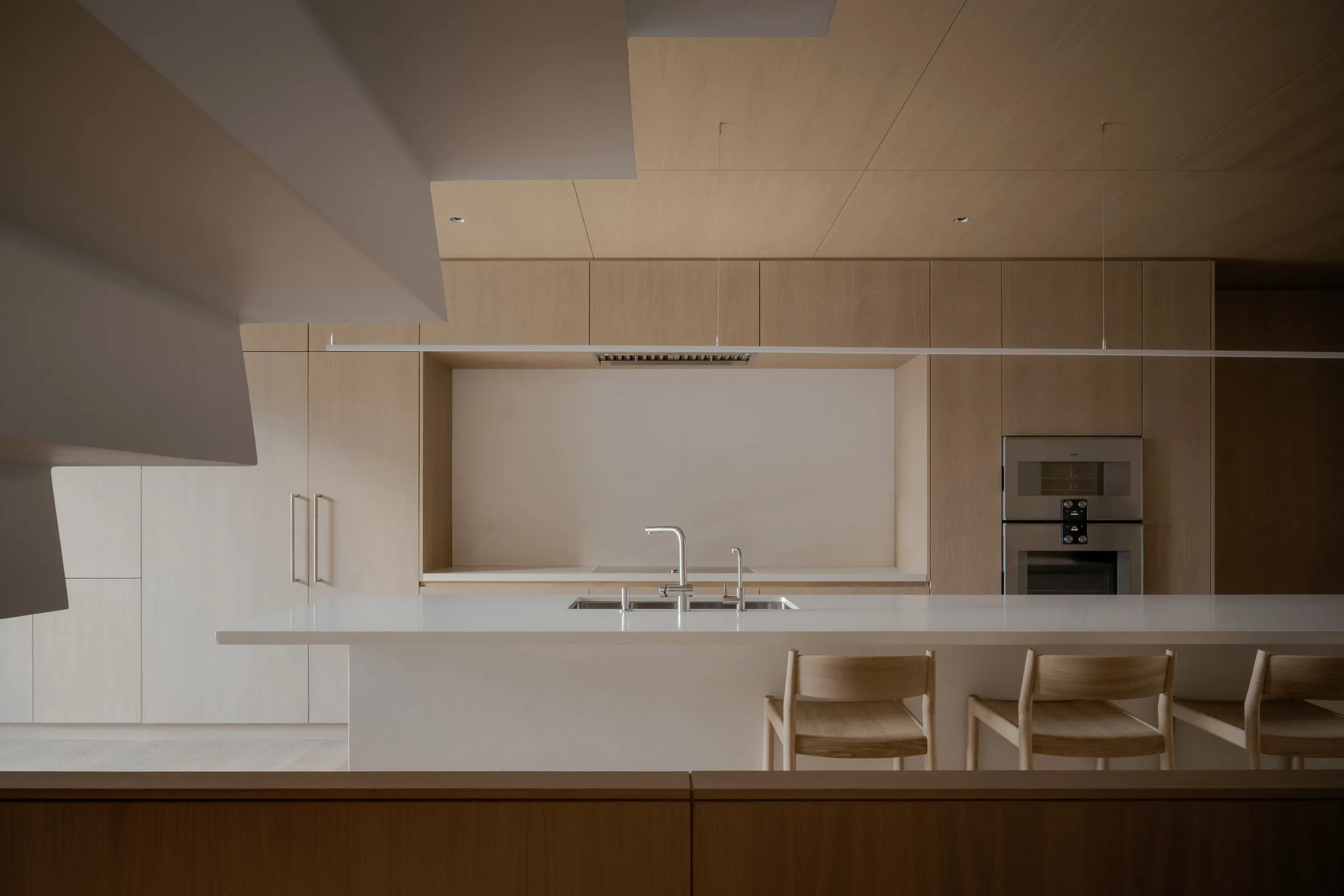 The kitchen at Bellwoods Residence, a white island and counter run below white oak uppers and an oak-lined backsplash niche, with wall ovens set into the run.