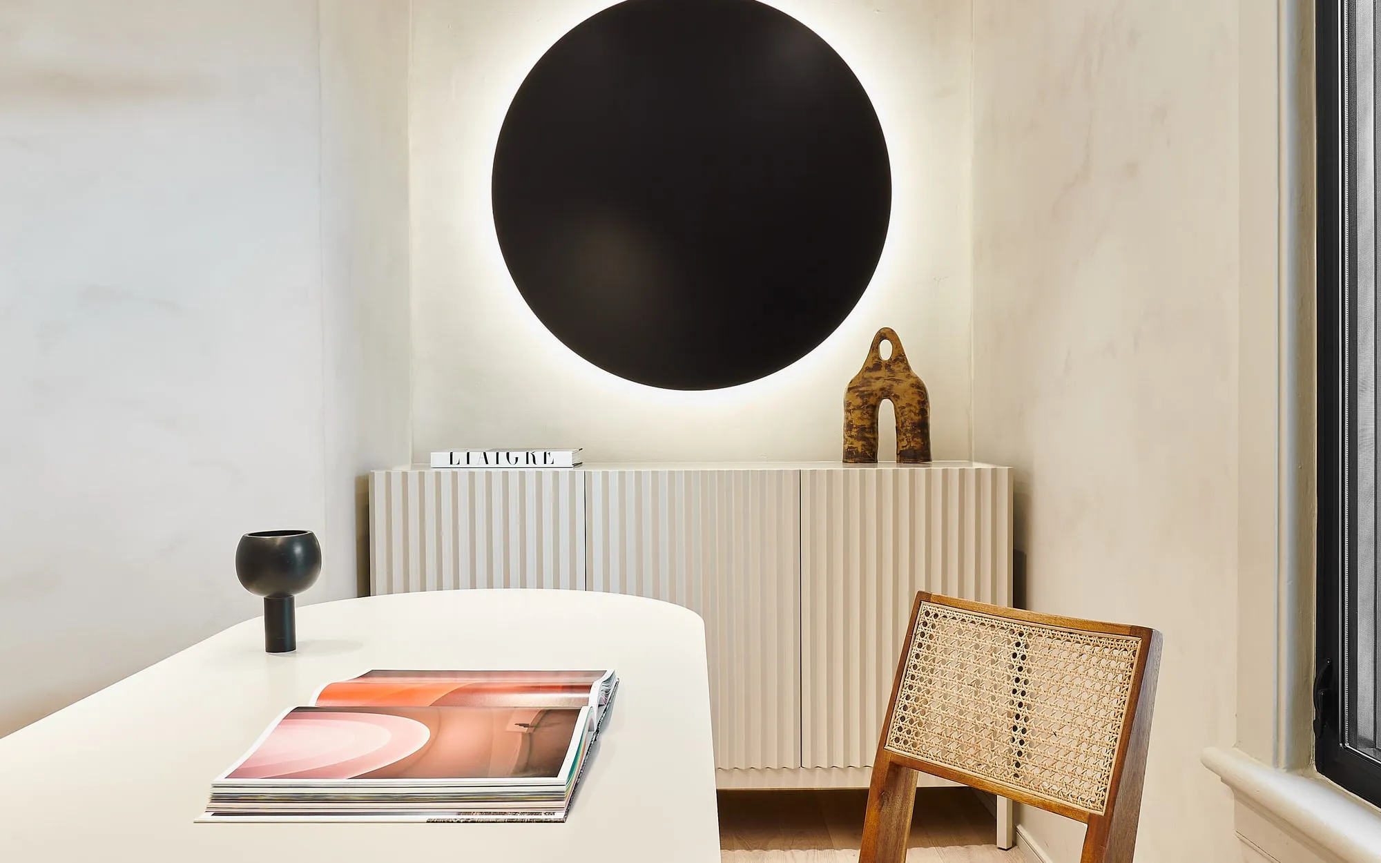 The Adelaide Project: a fluted cream credenza below a backlit black disc, with the rounded end of a white table and a cane chair in front, against plaster walls, Toronto.