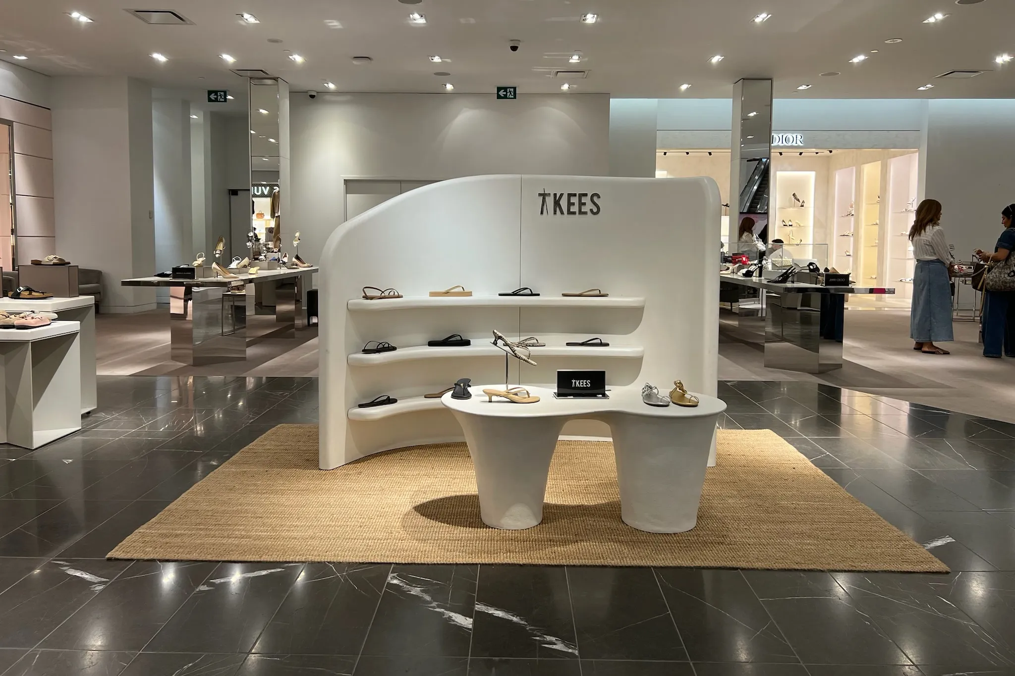 TKEES retail fixture at Holt Renfrew: a curved plaster-finished display wall with three shelves growing out of its face and a double-pedestal table in front, set on a jute rug on the shoe floor.